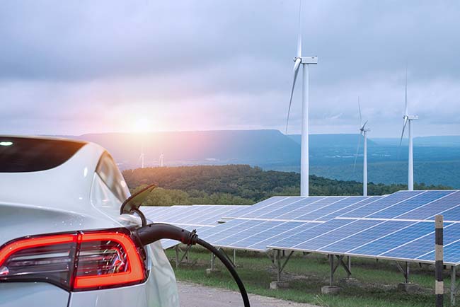 An electric vehicle plugged in and charging with solar panels and windmills in the background indicating a green energy source. 